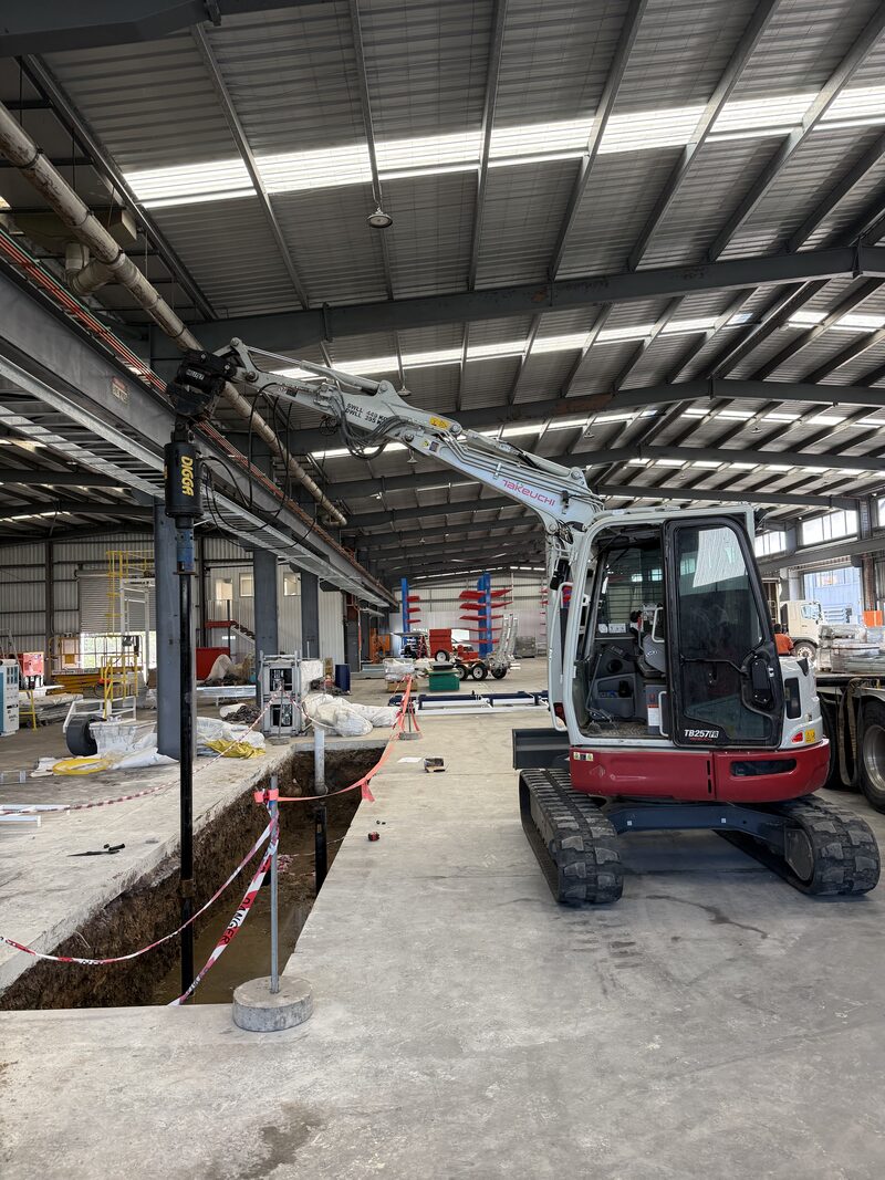 Excavator installing steel screw piles inside an operating industrial warehouse