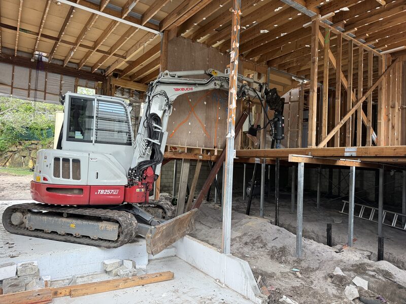 Excavator with hydraulic drive head installing steel screw piles beneath an existing home during an extension