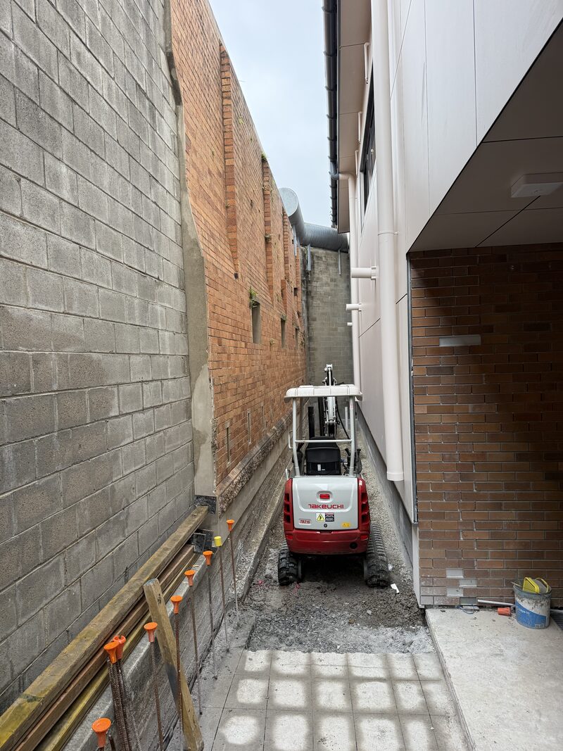 Mini excavator installing screw piles in a tight-access walkway at Boggo Road, Brisbane